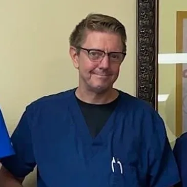 A man in blue scrubs smiles for the camera.